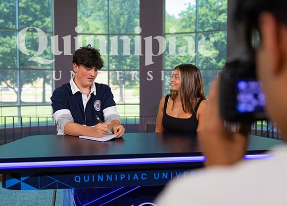Two students speaking at a newscast podium while another student films the discussion