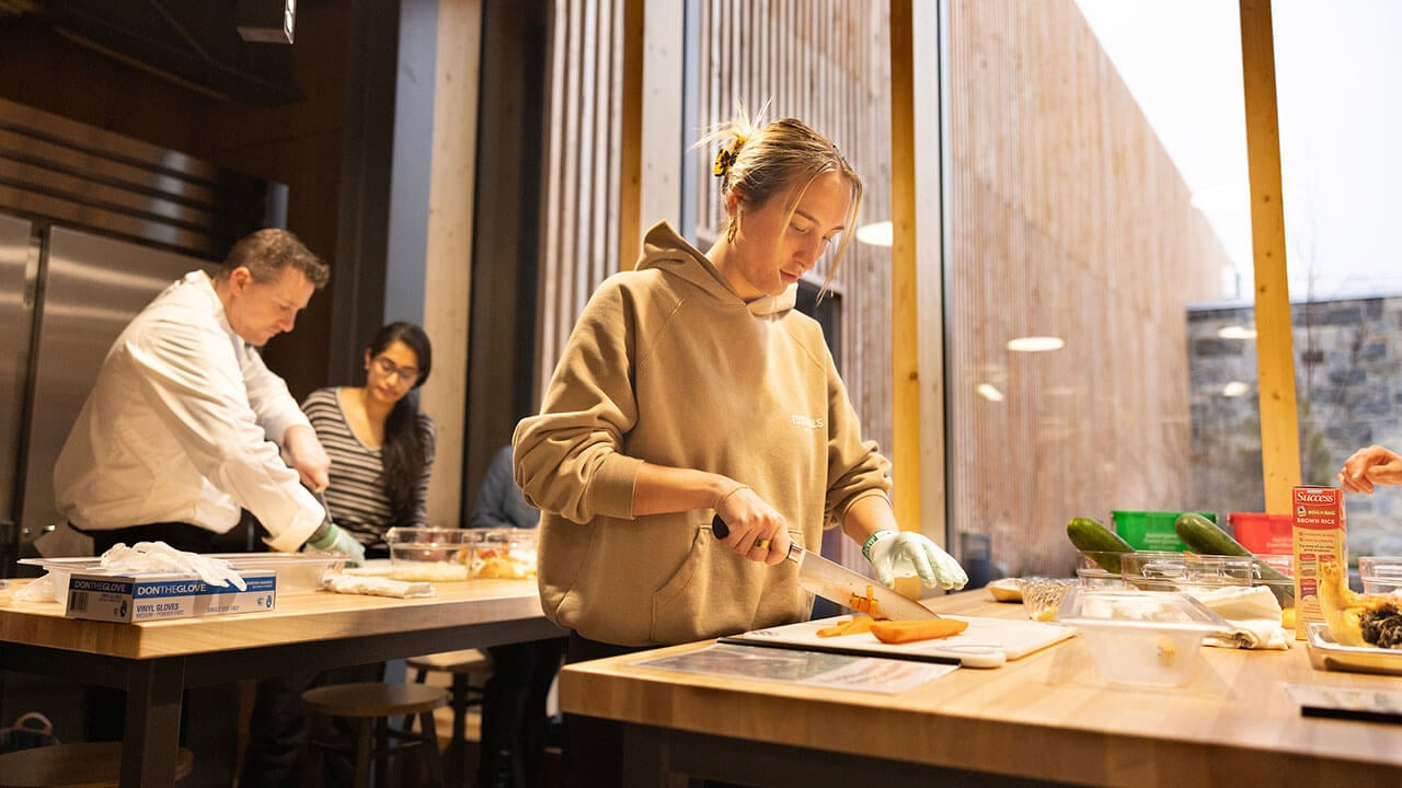 female student in a gray hoodie chops a sweet potato while in the background the chef helps another student