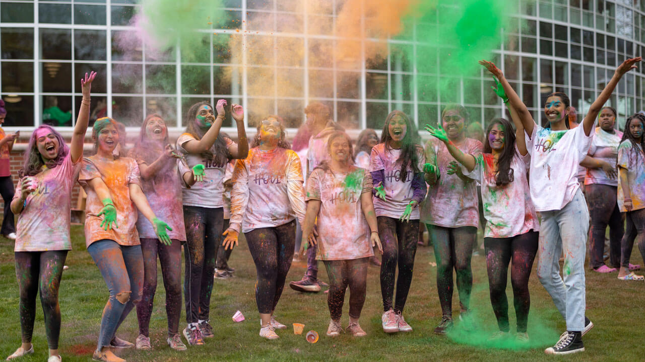 A large group of students throw colored powder in the air while celebrating Holi