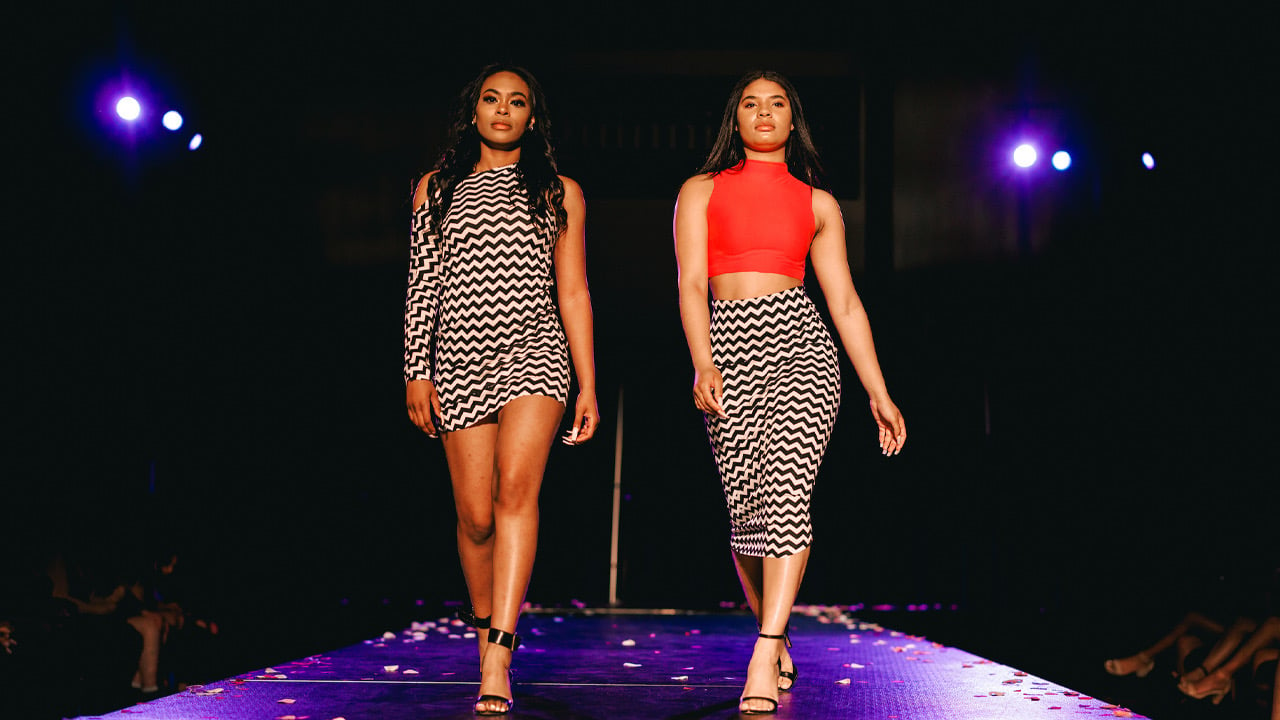 Two students in patterned dress and skirt walk the runway