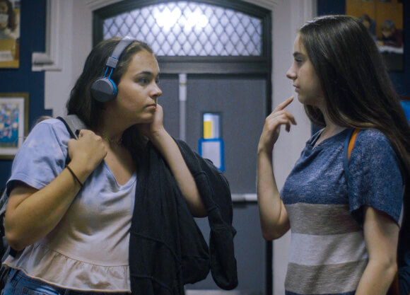 Two girls looking at eachother standing in a school hallway. The one on the left has brown hair and headphones. The one on the right has long brown hair.