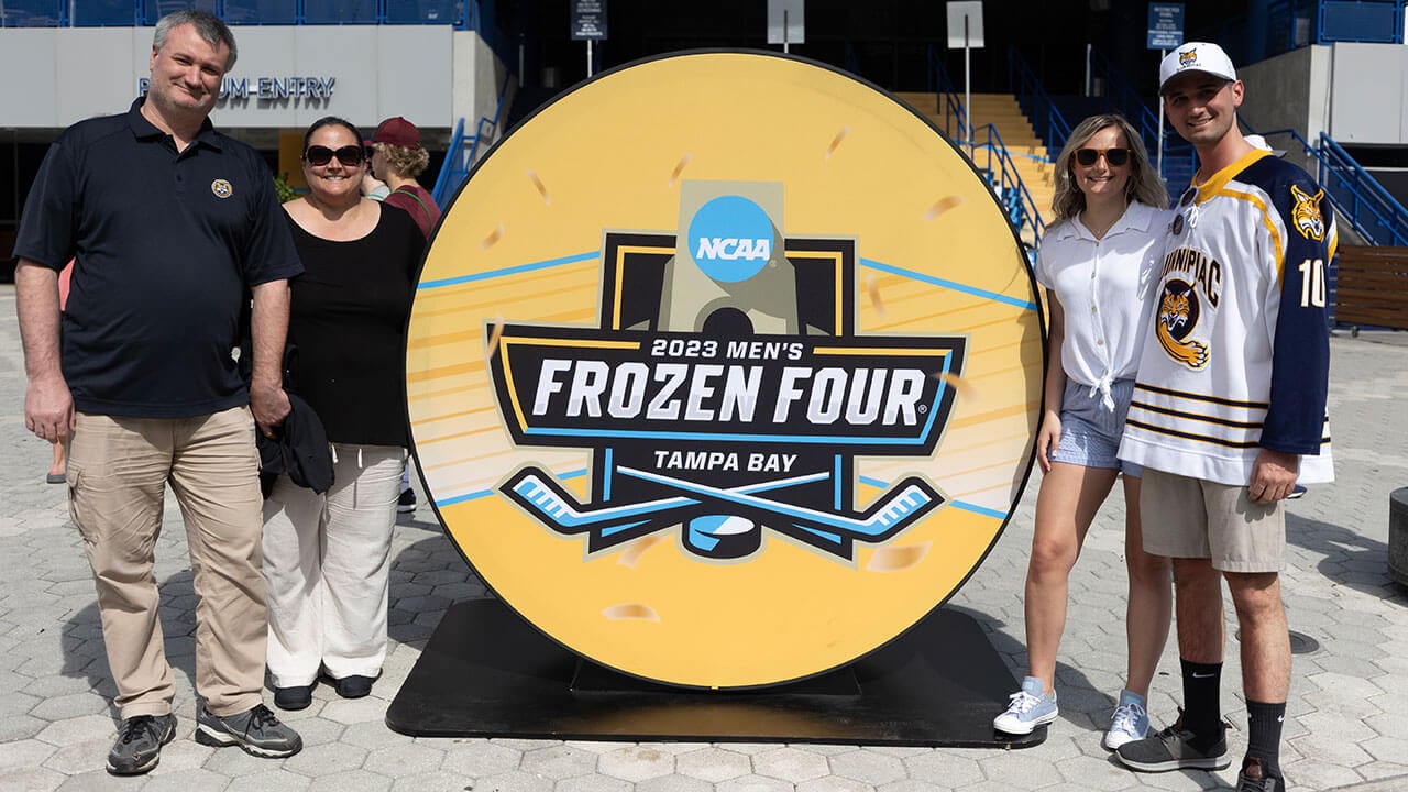 ryann glushek, a platinum blonde wearing a white shirt and light blue shorts, poses by a puck shaped frozen four in tampa bay sign with her brother alex in a quinnipiac hat and hockey jersey, and other the side are her parents