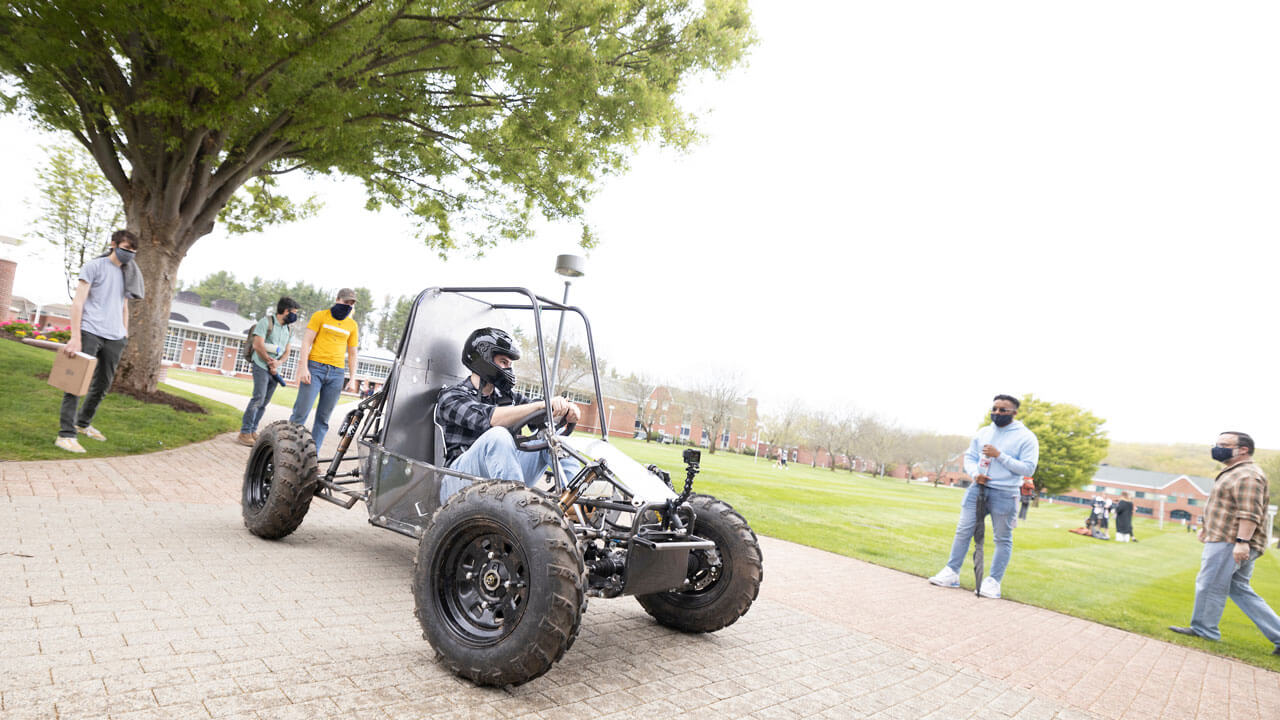Student sitting in baja vehicle on campus surrounded by a group of students.