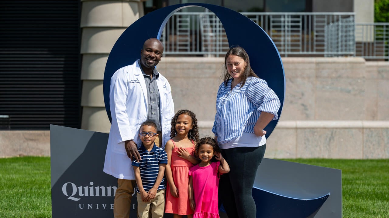 Family poses in front of Quinnipiac sign, smiling
