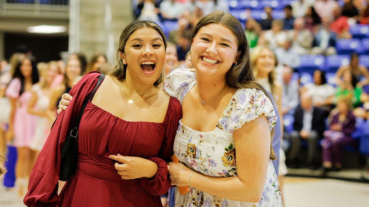 two female students, one in a wine red dress and one in white and lavender dress, both with brown hair, hug each other and smile for the camera