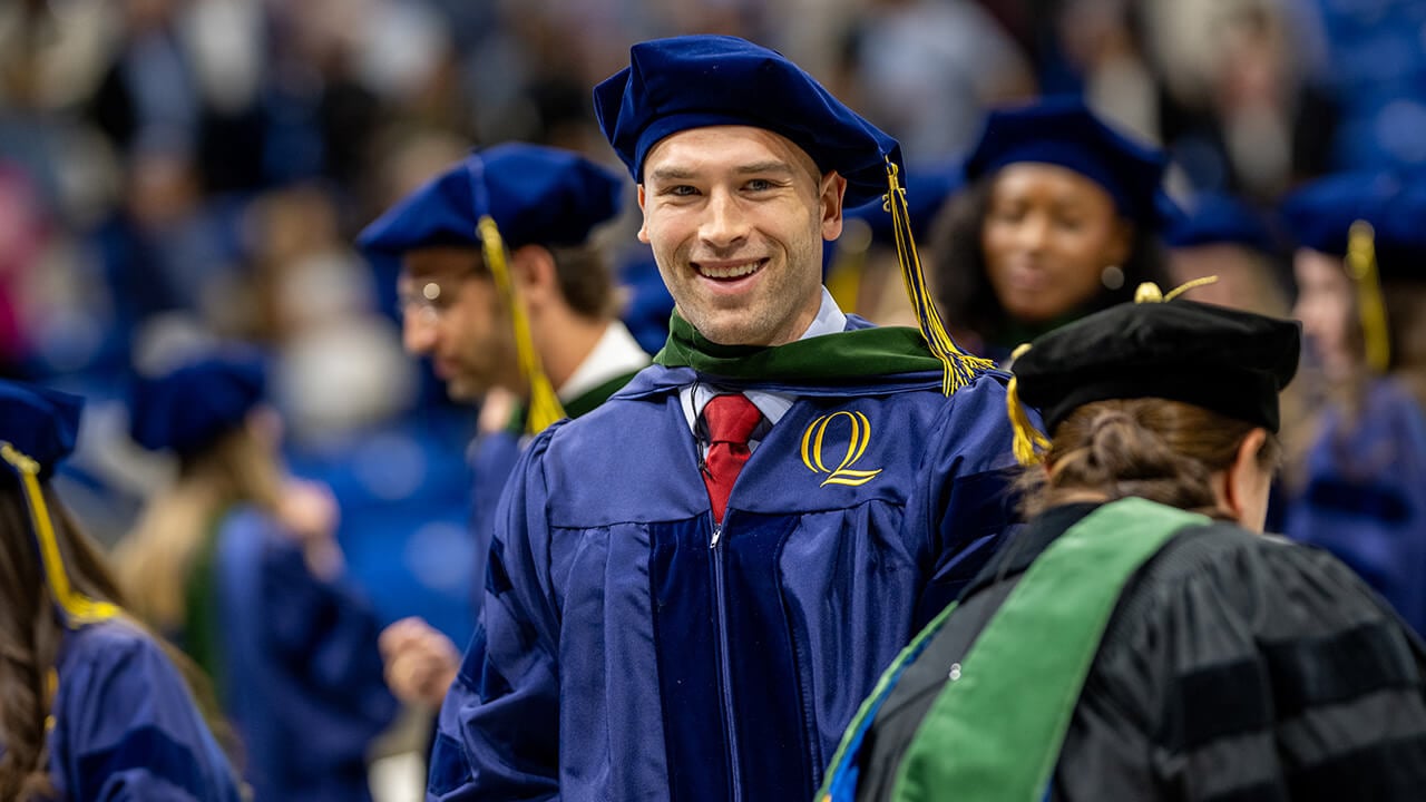 Graduate smiles during ceremony around peers, as they stand in the background