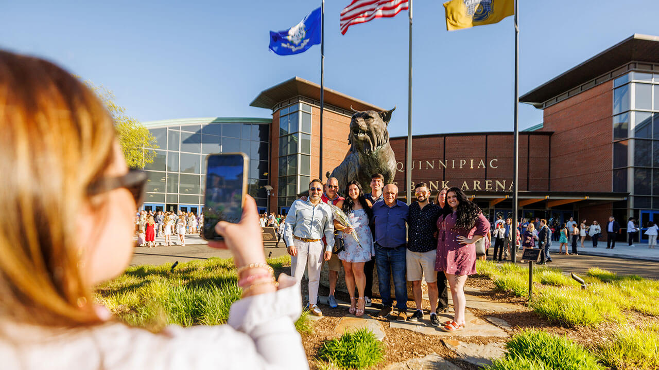 woman holds up her phone to take a picture of one of the pinning recipients and the people around her in front of the bobcat statue outside of the M&T Bank Arena