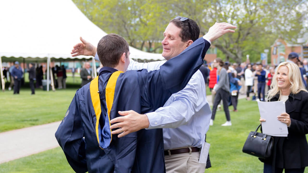 Graduate goes in for a hug with a family member while others smile while watching