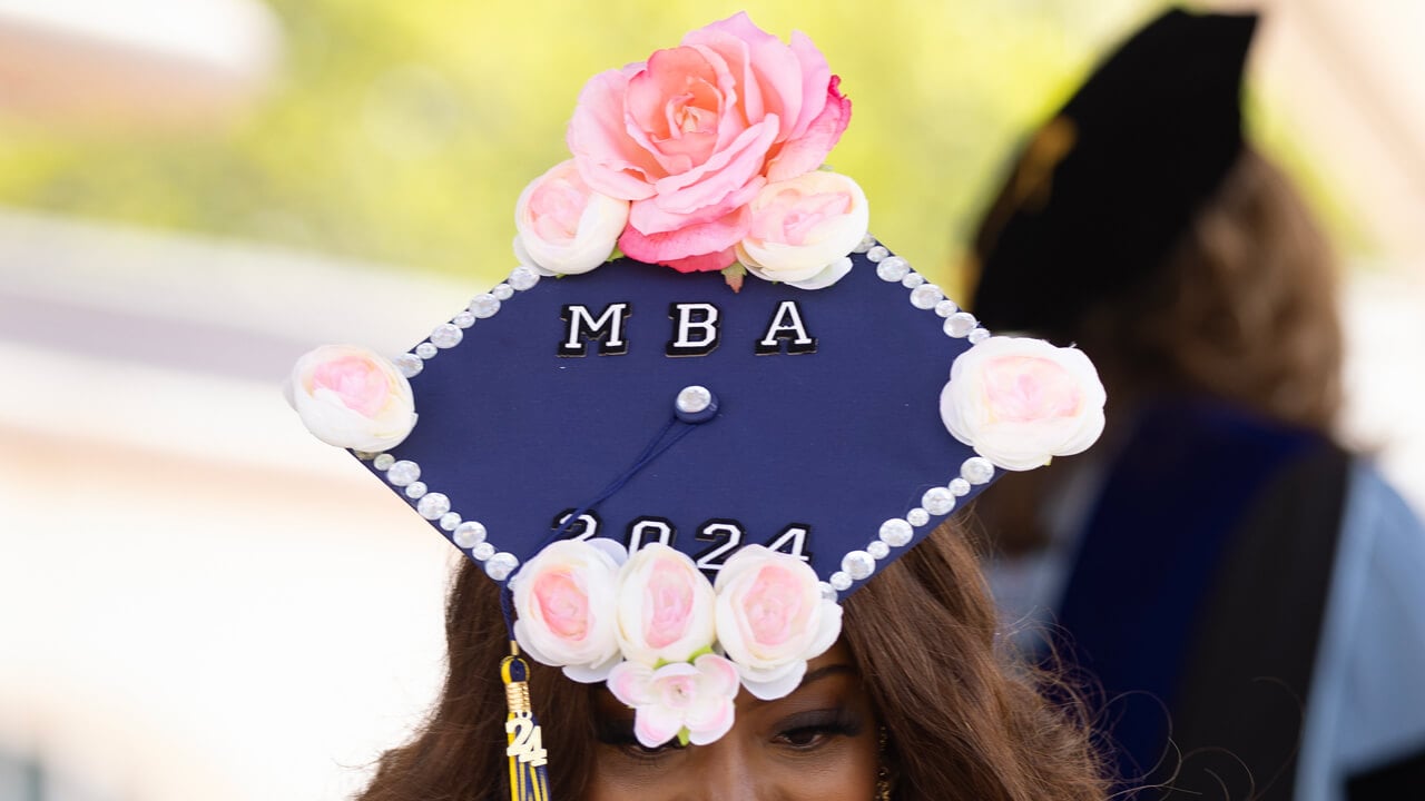 A graduate wears a graduation cap decorated with the flowers and words MBA 2024