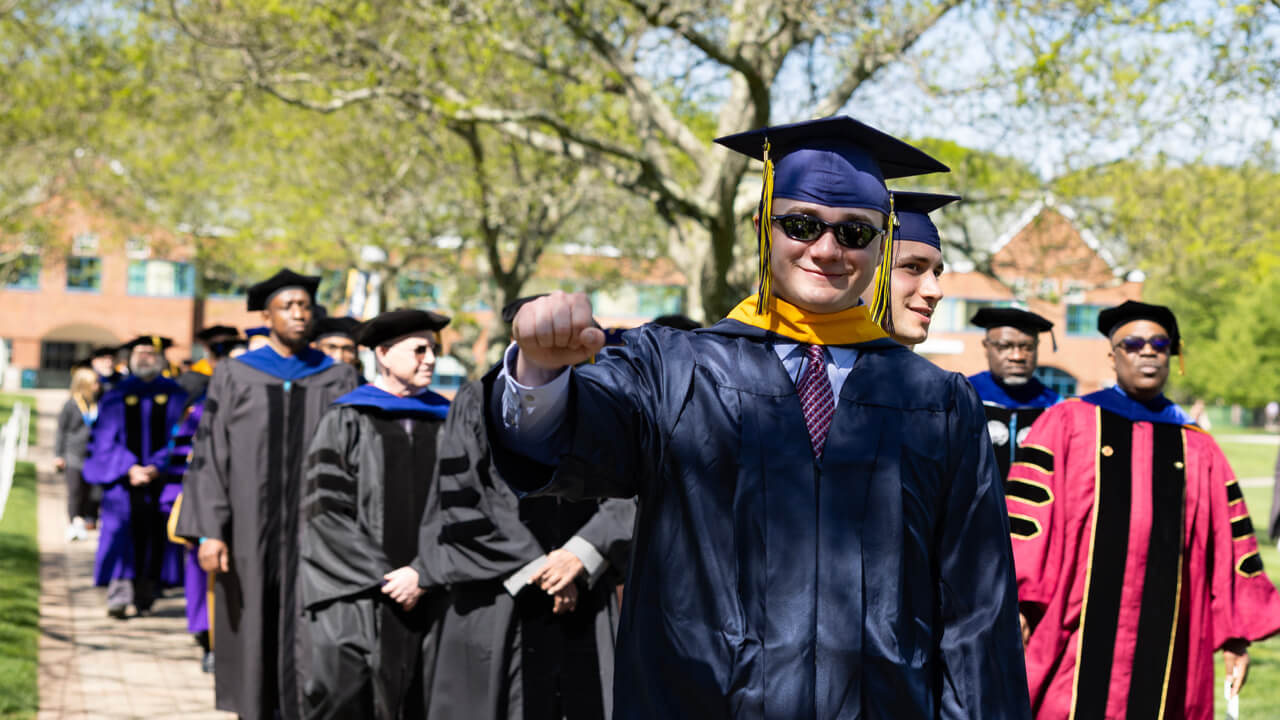 A graduate gives a fist bump into the air as he walks down the quad with faculty around him