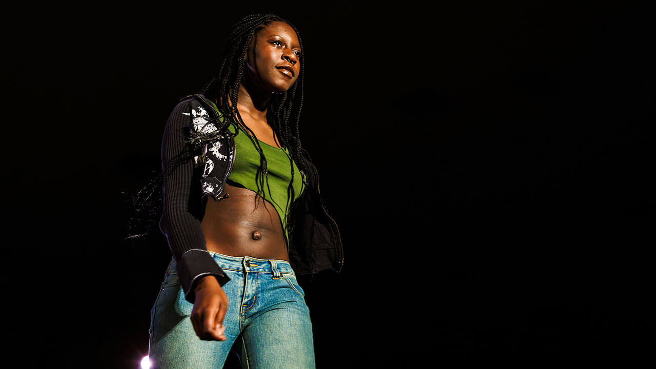 woman with braided hair and a green cropped tank top, black cropped sweater, and blue jeans walks down the runway