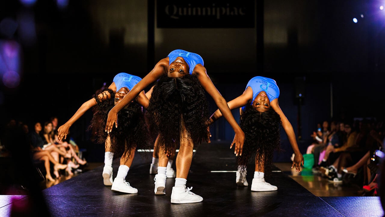three black women with long wavy hair in white tops bend over backwards while dancing
