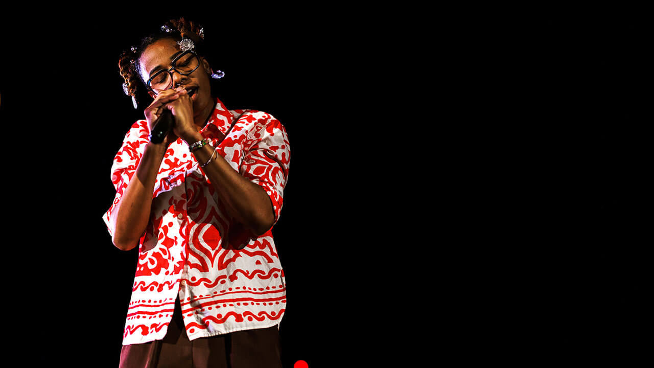 black woman with short braids and glasses wearing a red and white patterned shirt sings into the microphone with her eyes closed