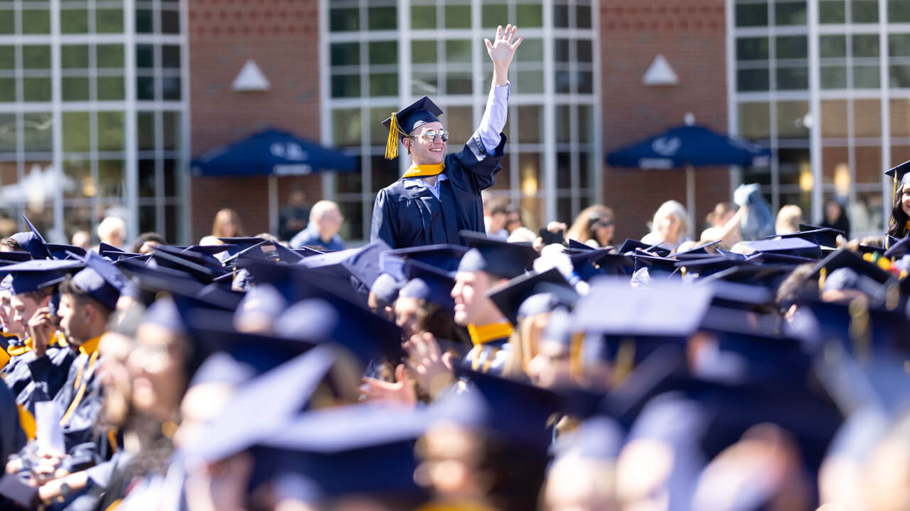 A business graduate in sunglasses stands and waves among a sea of seated graduates