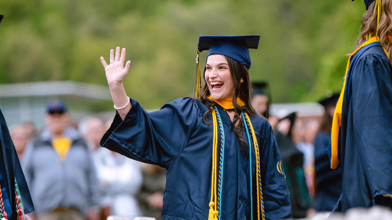 A graduate smiles broadly as she waves to family while she walks across the quad