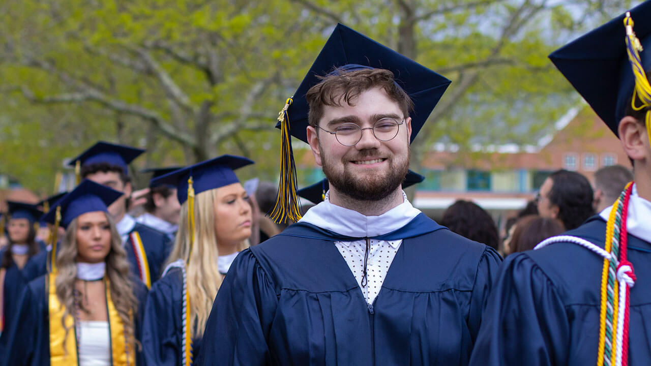 A graduate smiles as he walks across the quad with dozens of other graduates