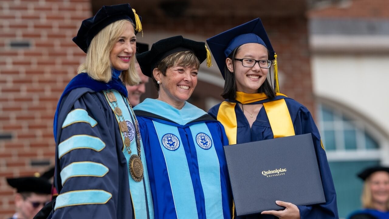 Graduate in cap and gown pose with photo with Marie Hardin and Monique Drucker