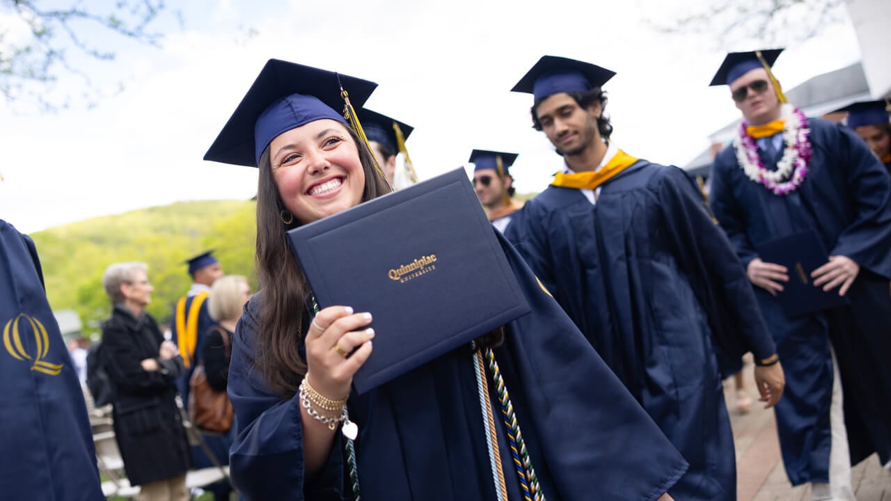 A graduate holds up her diploma and smiles as she walks down a quad path