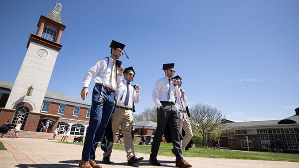 A group of students walk away from the library on Quinnipiac's Mount Carmel Campus Quad wearing graduation caps and carrying their gowns over their shoulders.