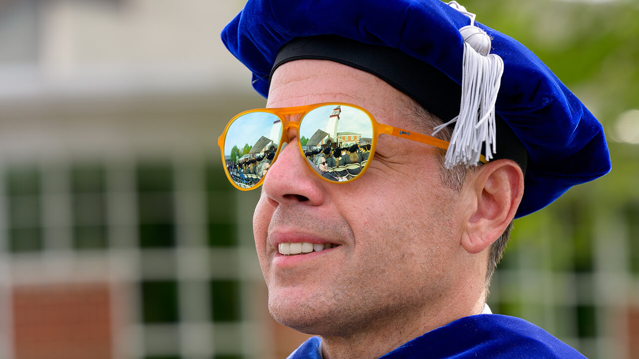 Reflection of the commencement and the Arnold Bernhard library are shown in orange reflective sunglasses on a graduate