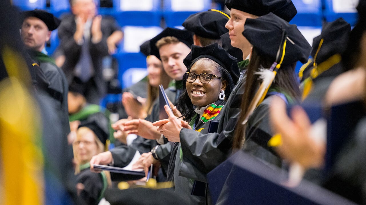 black female graduate smiles to the side while clapping in the crowd