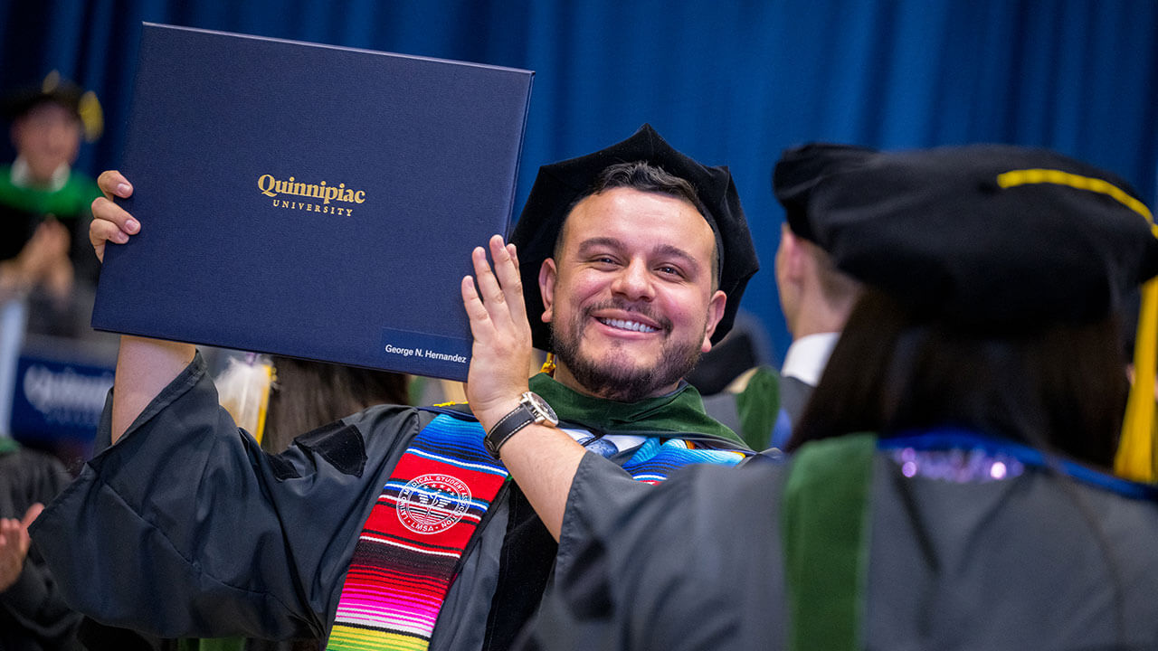 graduate wearing a latino medicinal society stole holds up his diploma