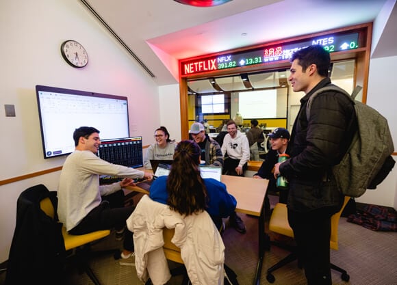 A group of students in a study room with a financial stock ticker in the background