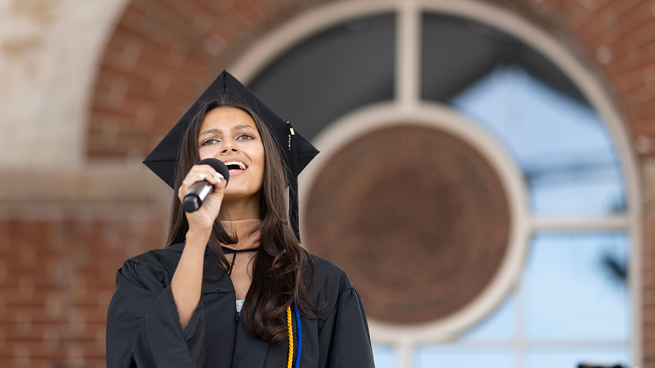 Graduate sings the national anthem in front of the Arnold Bernhard library