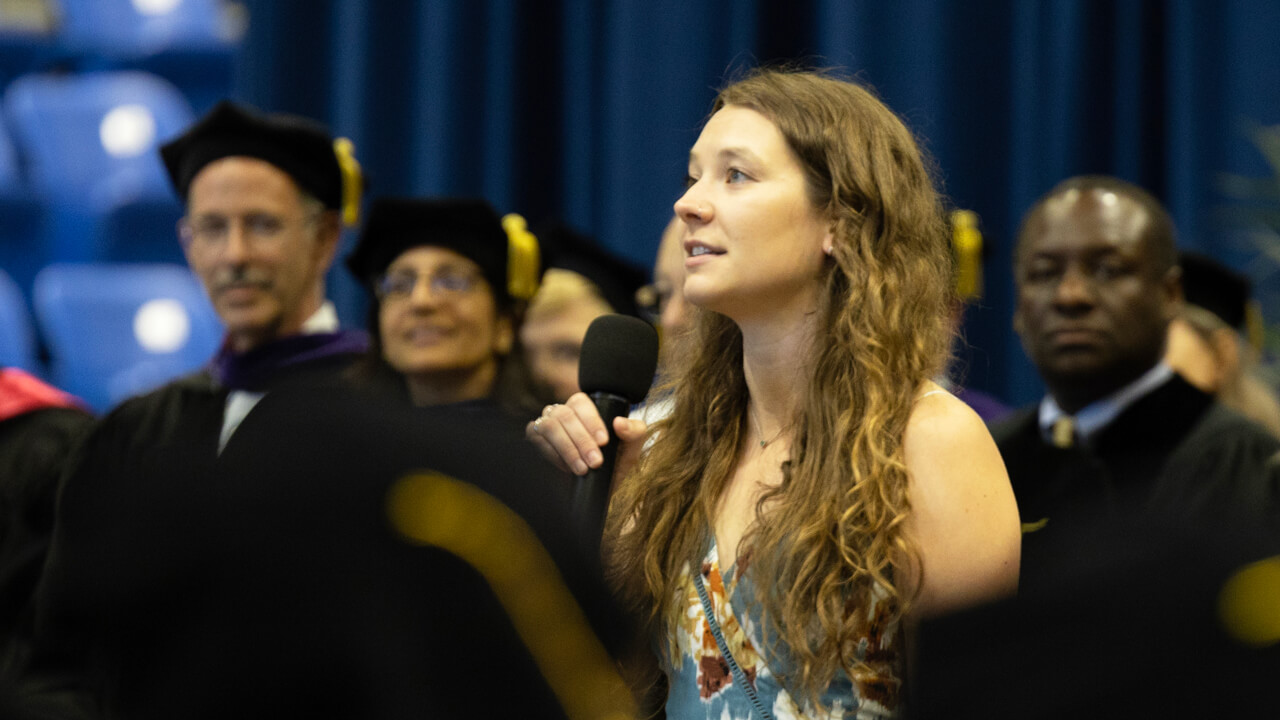 Cameron Chaplen holds a microphone and sings in front of the graduates