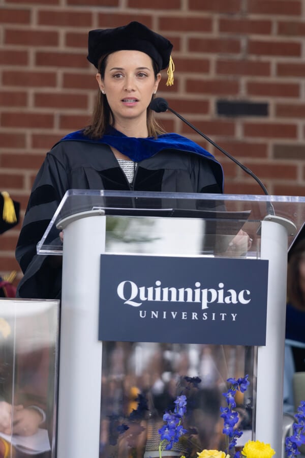 Jill Mayer wears commencement robes and speaks into a microphone