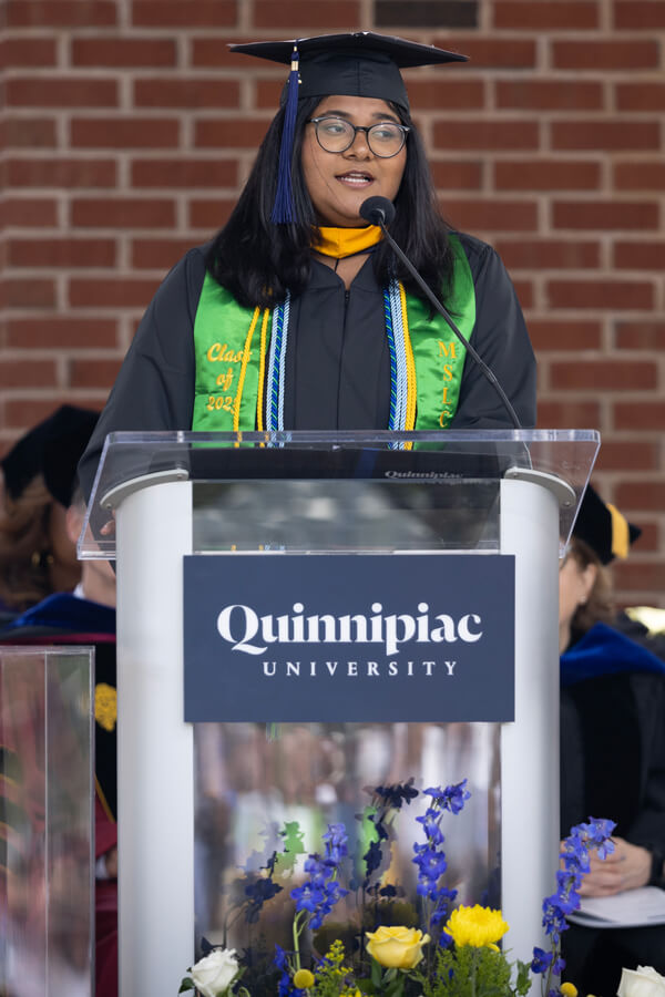 Hephzibah Rajan speaks at a Quinnipiac podium with flowers in front of it