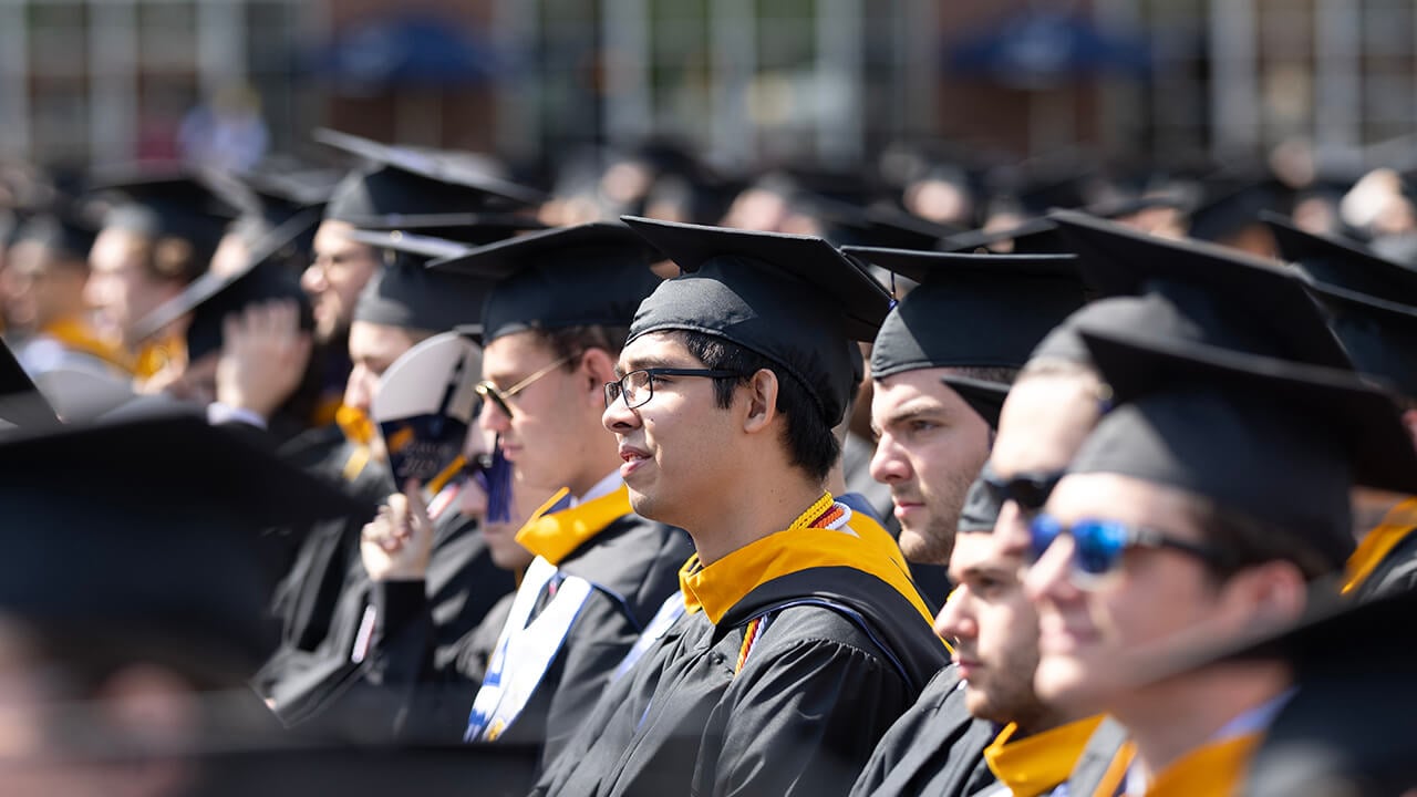 Graduate wearing glasses looks towards the stage while seated in the crowd