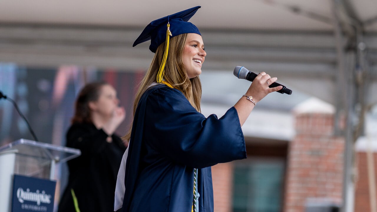 Hannah Jack in her cap and gown holds a microphone and sings on the library steps