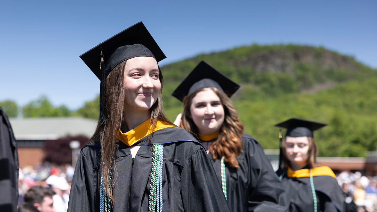 Several graduates walk across the quad with Sleeping Giant State Park in the background