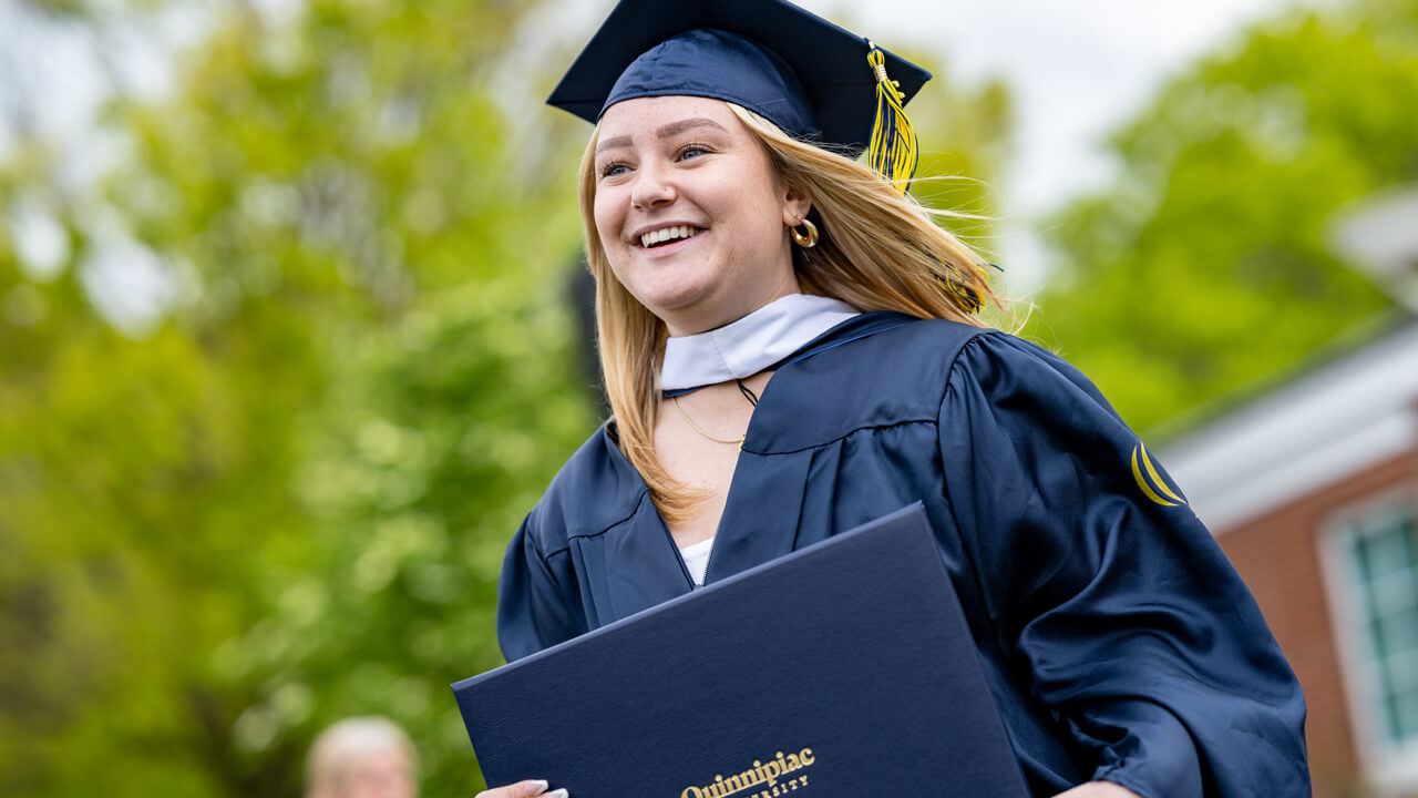 A graduate smiles while holding her Quinnipiac diploma as a breeze blows her hair
