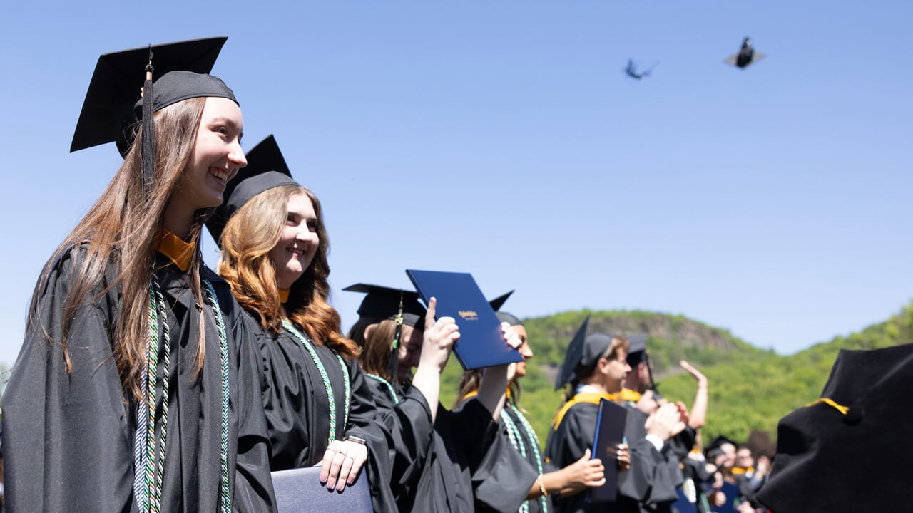 Graduates smile, wave and toss caps into the air in front of a blue sky
