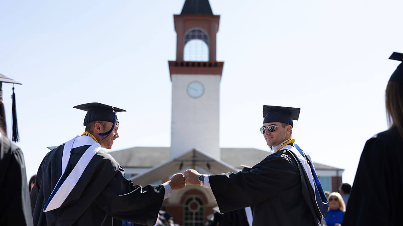 two graduates fist bump in front of the arnold bernhard library in their caps and gown