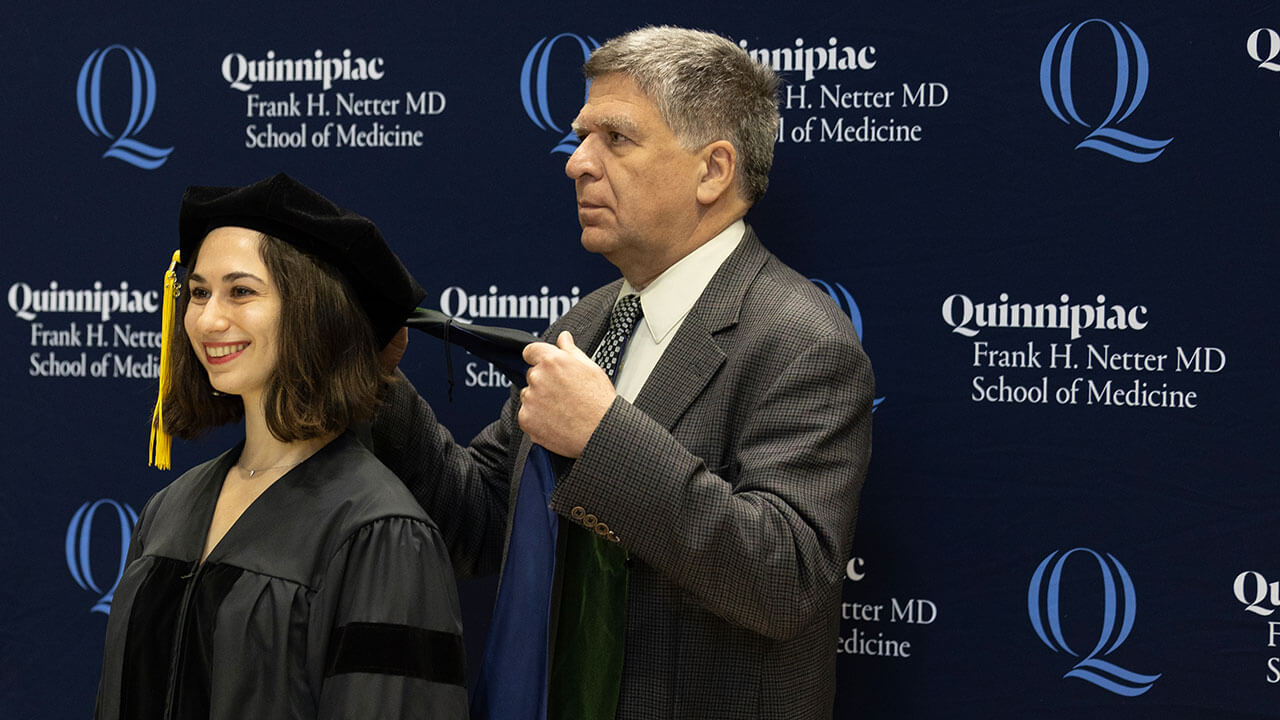 school of medicine graduate with short dark hair receives a green hood by a gray haired man in a gray suit