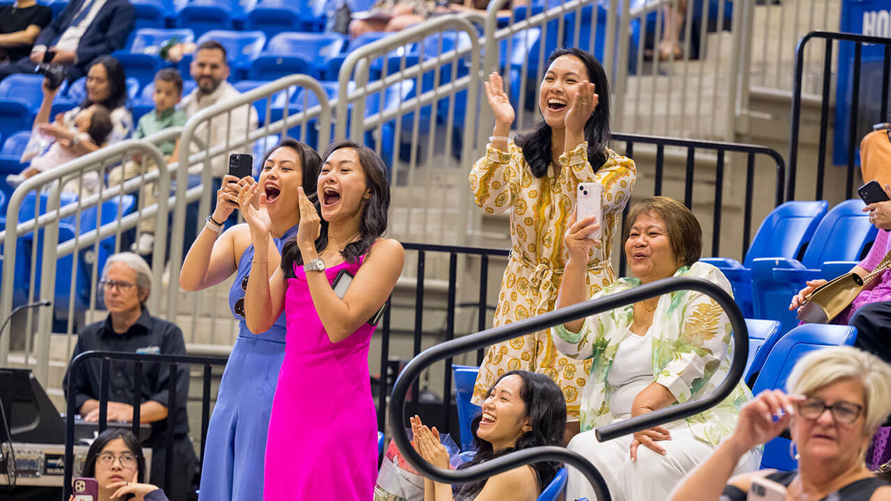three young women wearing bright colors cheer and clap for the graduates