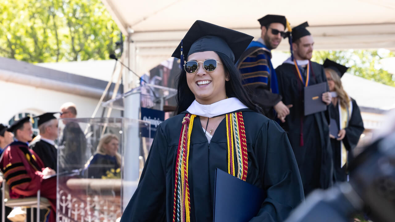 A graduate wears sunglasses and smiles broadly as she walks down the library steps with her diploma