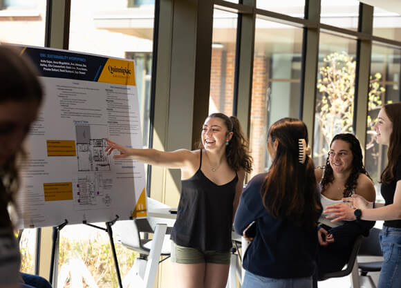 A student in The Grove points to a poster presentation board while other students look on