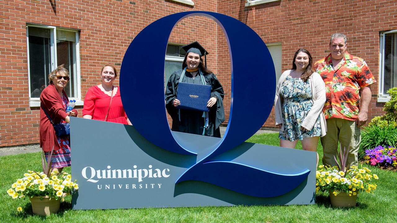A graduate and their family standing with a Quinnipiac University sign on the lawn on Bobcat Way.