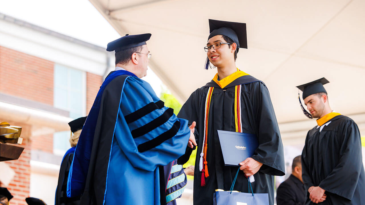 male graduate wearing glasses shakes the dean's hand while holding his diploma