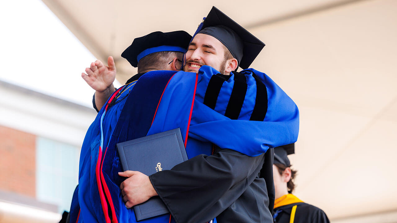 male graduate grins broadly while hugging professor with his diploma