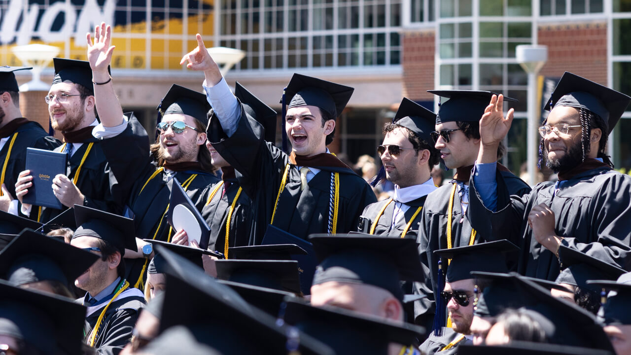 A row of graduates stand and cheer during a Quinnipiac Commencement ceremony