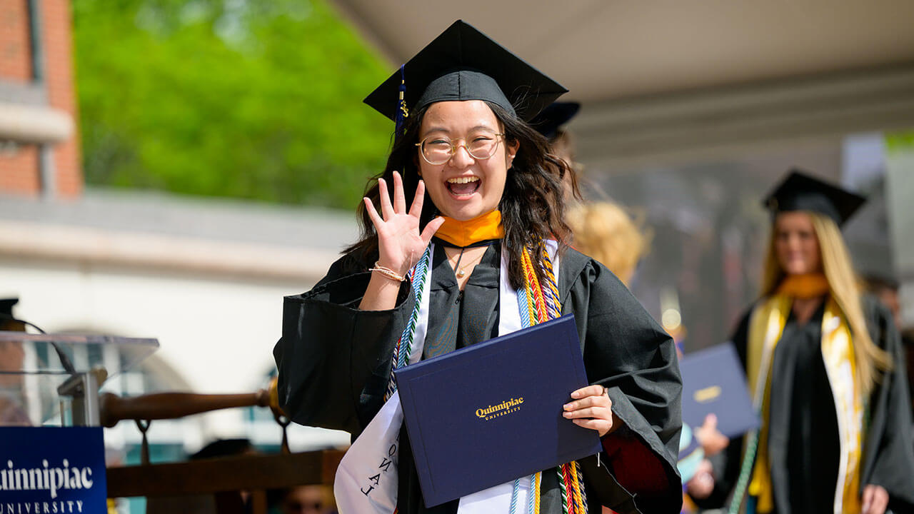 female graduate smiles and waves to the camera from the stage