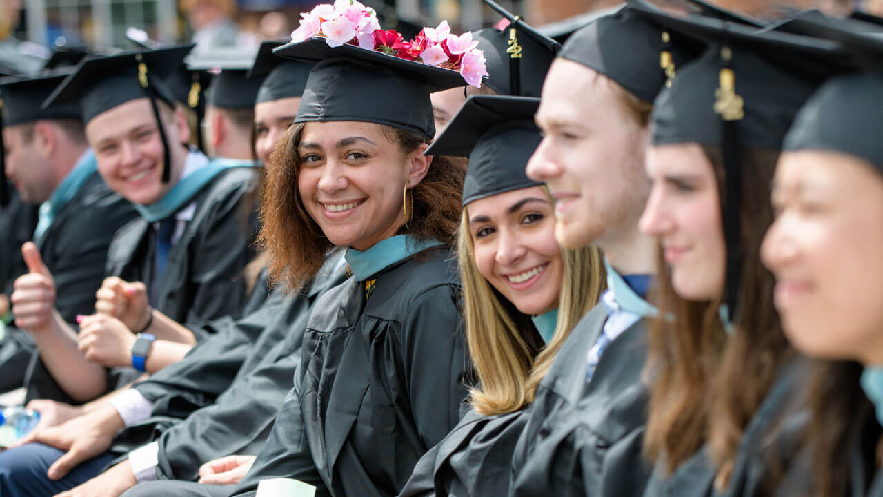 Graduating graduate student with pink floral cap smiles while sitting with other students at ceremony