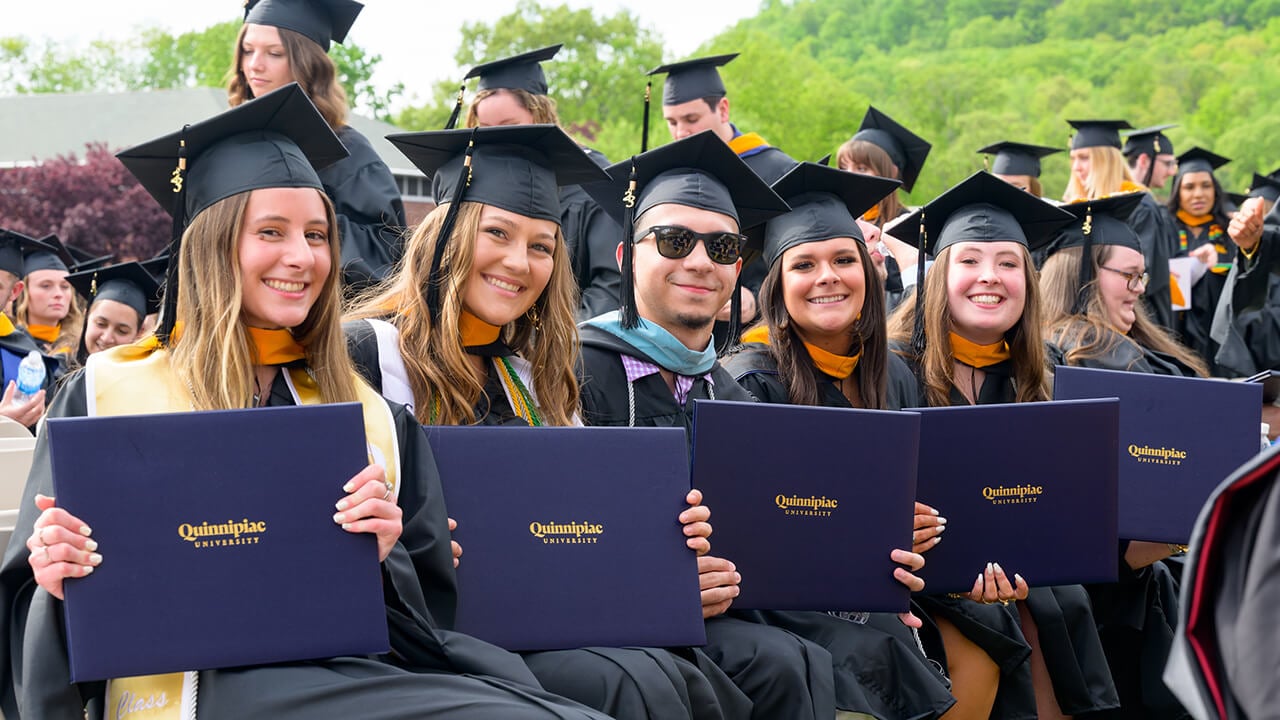 A row of seated graduates hold up their diplomas and smile together