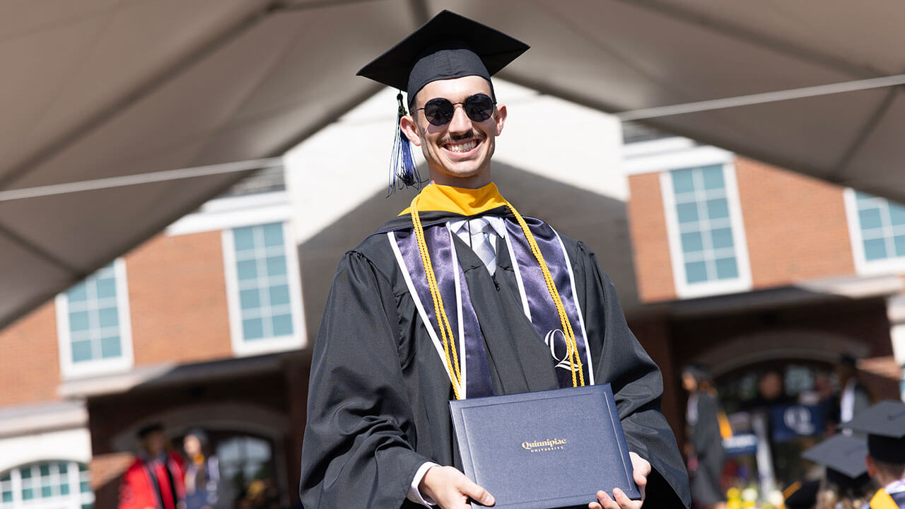 graduate wearing sunglasses poses in the sunlight with his diploma