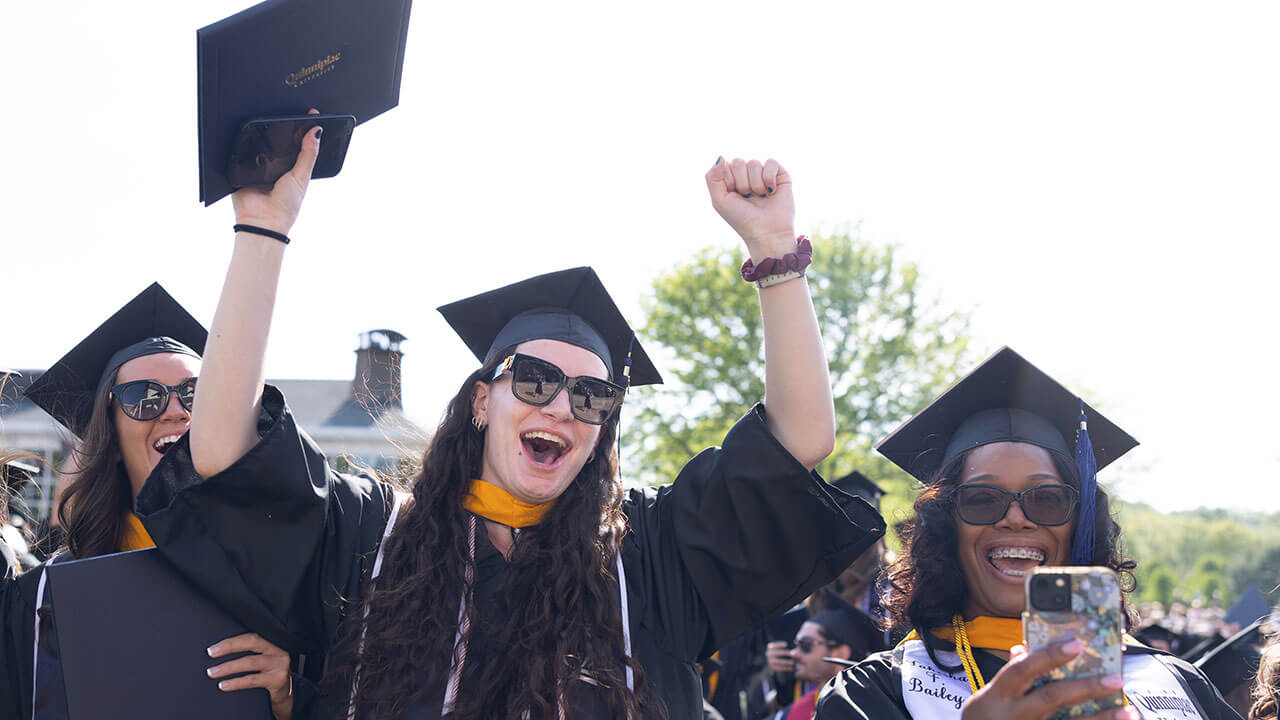 A graduate throws her hands in the air in celebration while another takes a photo
